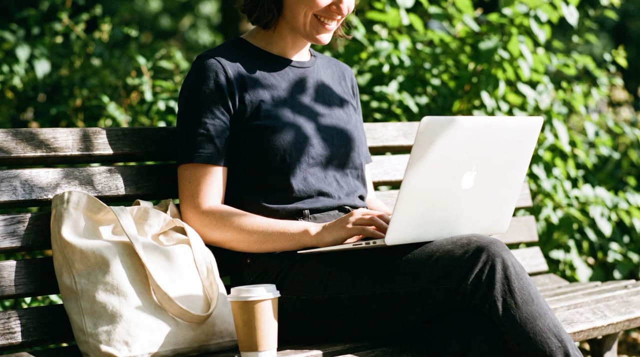 An open laptop on a park bench under leafy trees, with sunlight dappling the keyboard and path behind it